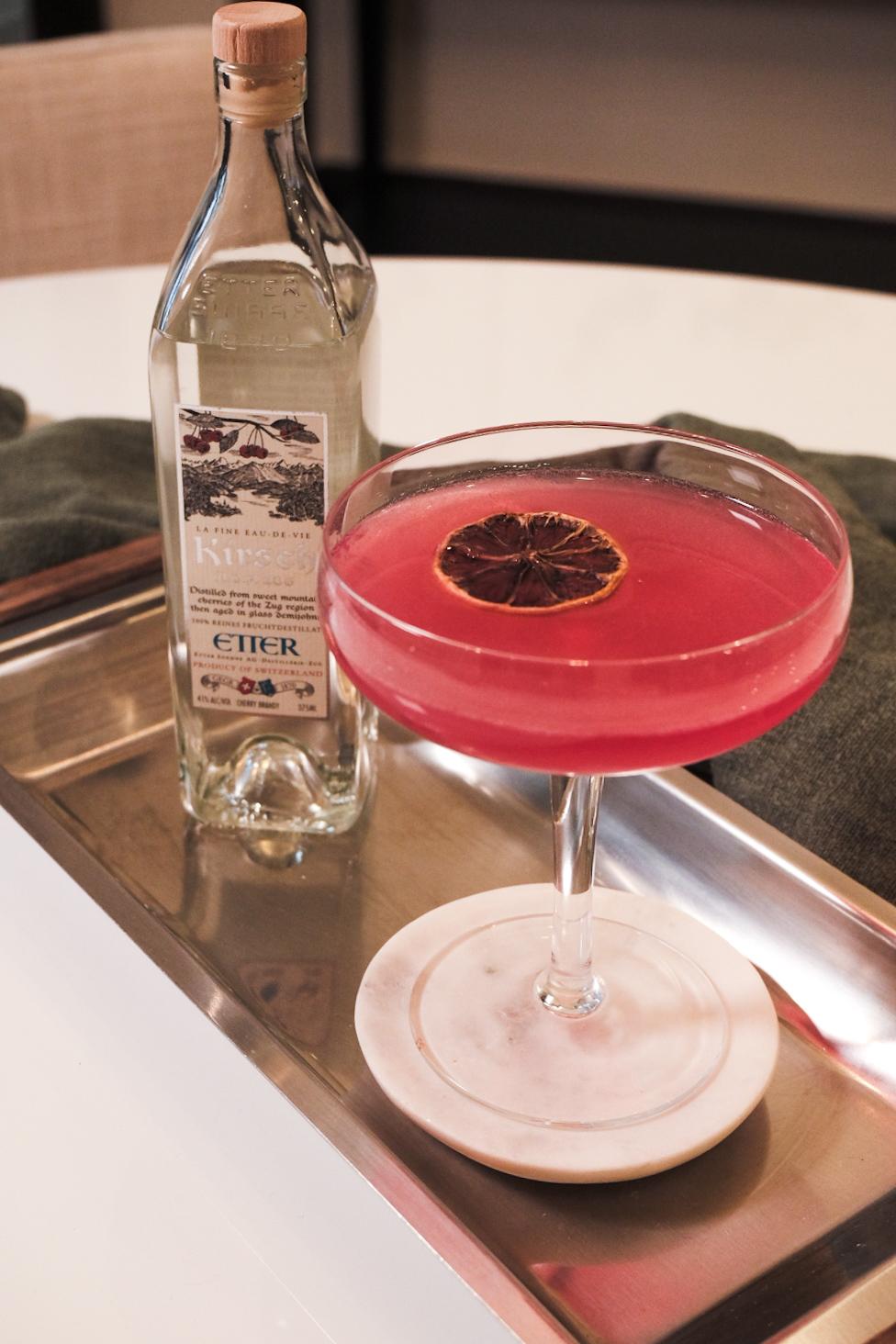 An example of the Kirsch Cosmo, the mixed drink (drink), by Toby Cecchini, NYC and Hannah Lanfear, London, featuring Etter Kirsch, vodka, orange-flavored liqueur, lime juice, cranberry juice, and lemon wheel